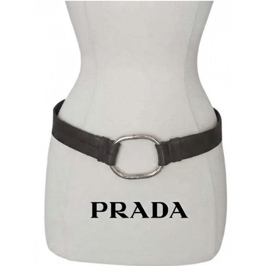 A black leather belt with a large silver-tone oval buckle displayed on a white mannequin torso. The word 'PRADA' is printed in black text below the mannequin.