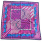 A square silk scarf with a vibrant pink, blue, and white geometric pattern featuring stylized figures, laid flat on a dark wood floor.