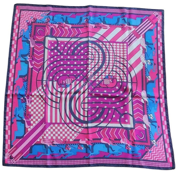 A square silk scarf with a vibrant pink, blue, and white geometric pattern featuring stylized figures, laid flat on a dark wood floor.